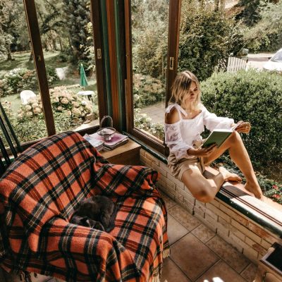 Woman reading by a sunlit window, with a cat sleeping on a tartan blanket-covered chair nearby.