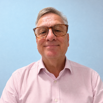 Smiling older man with glasses in a light pink shirt, standing in front of a pale blue background.