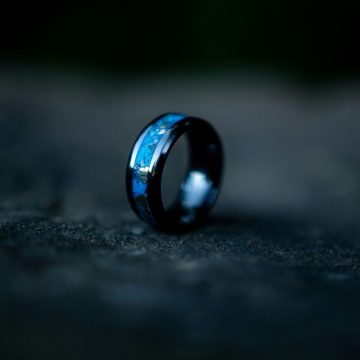 A black ring with a blue inlay rests on a dark, textured surface with a blurred background.