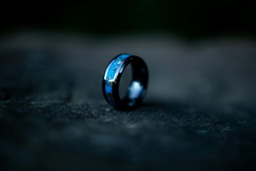 A black ring with a blue inlay rests on a dark, textured surface with a blurred background.